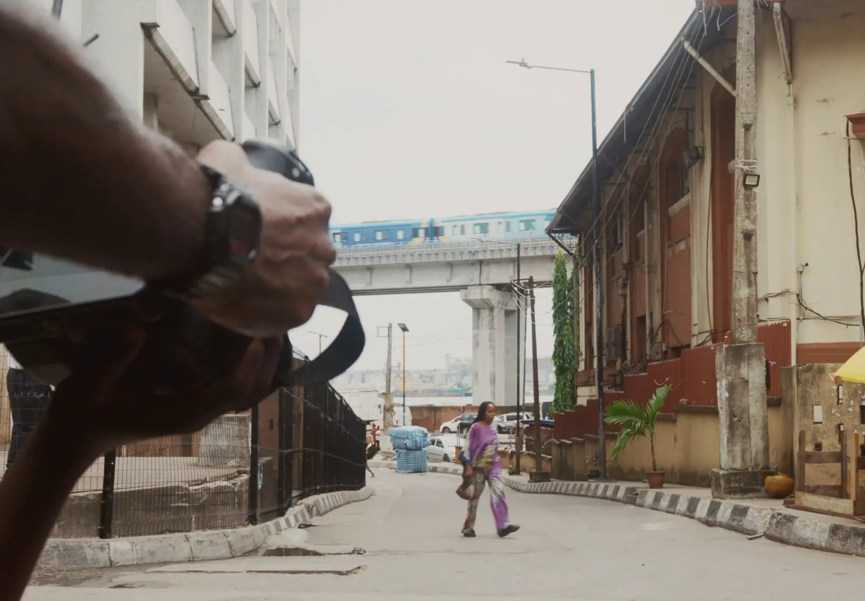 A camera operator filming a woman walking down a Lagos street under a passing train